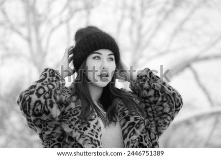 Portrait of young adult woman with long hair and plump lips, wearing fur coat with leopard print and knitted hat. Woman is looking away while raising hands and straightening hat. Black and white