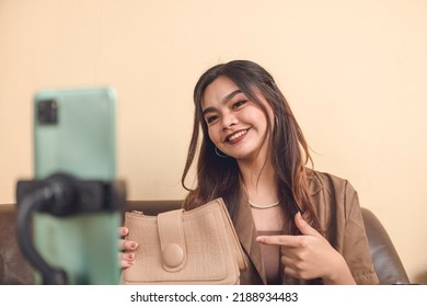 Portrait Of A Young Adult Woman Taking A Selfie. Live Selling Her Handbag Using Her Phone Attached To A Stand. A Side Hustle Using Social Media To Promote Her Products.