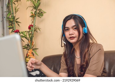 Portrait Of A Young Adult Woman Working Online Wearing Headset And Using Laptop Wearing Brown Smart Casual Attire.