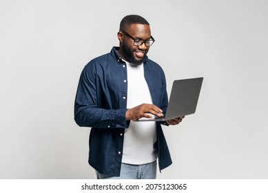 Portrait of young African American guy standing isolated on grey wall holding laptop, typing messages, chatting with friend on social media online, watching video, enjoying new application, websurfing