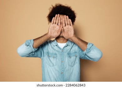 Portrait of young african american guy introvert in denim shirt standing with hands covering his face, hiding from people and society, escaping from reality. Body language, signs, gesture