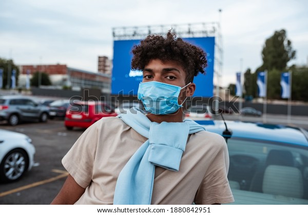 Portrait of young african american man in\
protective mask standing on car cinema screen background during\
coronavirus pandemic and looking at camera. Free time, safety and\
entertainment concept