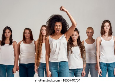 Portrait of young african american woman in white shirt and denim jeans raised her fist while looking angry at camera. Diverse women standing over grey background. Femininity concept. Selective focus