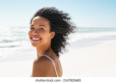 Portrait of young african american woman with curly hair at beach. Smiling black woman walking at the seaside while looking at camera. Happy carefree girl at sea.