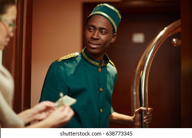 Portrait Of Young African Boy Working As Bellhop In Luxury Hotel, Getting Tip From Woman Guest For Delivering Luggage To Room