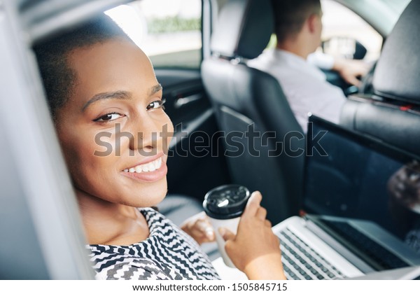 Portrait of\
young African businesswoman smiling at camera while working with\
laptop and drinking coffee during trip by\
car