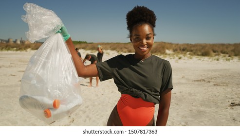 Portrait of an young african female volunteer is smiling in camera satisfied with picking up a plastic litter on a beach with a sea to protect an environment.