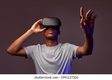 Portrait of young african teen boy in VR helmet looks away at copy space, isolated in studio, guy holds out hand forward. Gamer playing VR games on colourful background, VR gaming. - Shutterstock ID 1991127326