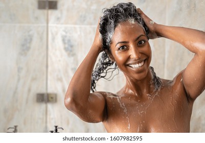 Portrait Of Young African Woman In Shower Washing Hair With Shampoo And Conditioner At Spa. Happy Black Woman Massaging Hair With Shampoo In Shower And Looking At Camera. Girl Washing With Copy Space.