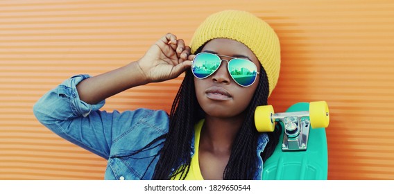 Portrait of young african woman with skateboard wearing a sunglasses, hat in casual over orange wall background - Powered by Shutterstock