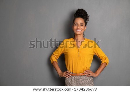 Portrait of young african woman standing with hands on waist and looking at camera. Confident stylish latin girl smiling isolated against grey background. Beautiful woman with copy space on gray wall.