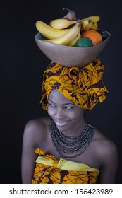 Portrait Of A Young African Woman In Traditional Dress With A Bowl Of Fruit On Her Head. 