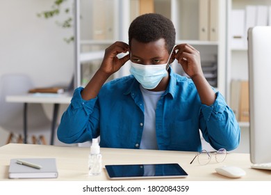 Portrait Of Young African-American Man Putting On Face Mask While Working At Desk In Post Pandemic Office