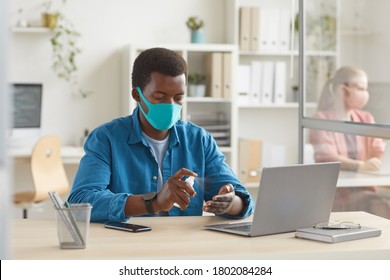 Portrait Of Young African-American Man Wearing Mask Sanitizing Hands While Sitting At Desk In Cubicle At Post Pandemic Office, Copy Space