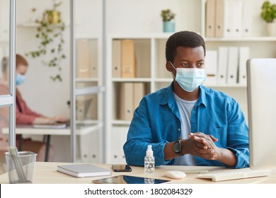 Portrait Of Young African-American Man Wearing Face Mask And Sanitizing Hands While Working In Cubicle At Post Pandemic Office, Copy Space