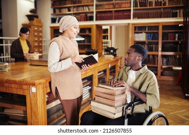 Portrait of young African-American man in wheelchair holding stack of books and chatting with friend at college library