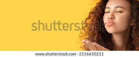 Portrait of young African-American woman with beautiful eyeshadows blowing air kiss on yellow background with space for text