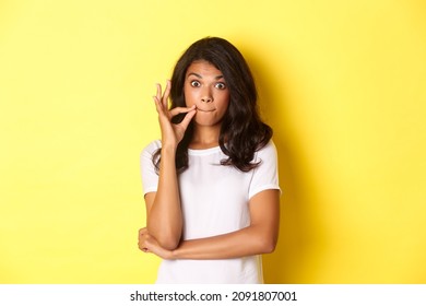 Portrait of young african-american woman making promise to keep secret, seal lips, zipping mouth with fingers, standing over yellow background