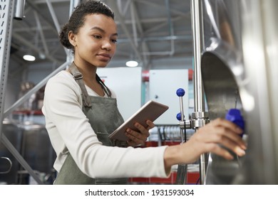 Portrait of young African-American woman operating brewing equipment at beer making factory and using digital tablet, copy space