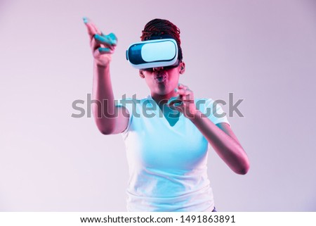 Portrait of young african-american woman's playing in VR-glasses in neon light on gradient background. Concept of human emotions, facial expression, modern gadgets and technologies. Pointing, shooting