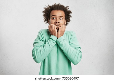 Portrait of young Afro American man with scared and anxious expression, bites finger nails, wears green sweater, being afraid of visiting doctor. Human facial expressions and negative emotions concept