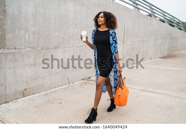 Portrait of young afro american woman walking while holding a cup of coffee and a bag outdoors.