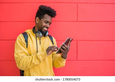 Portrait of young afro man using his digital tablet with earphones against red wall. Technology and urban concept. - Shutterstock ID 1780054073
