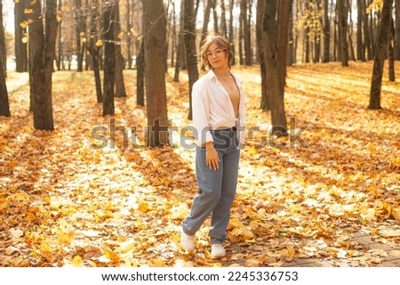 Portrait of young amazing teenage girl wearing jeans, shirt, glasses, standing among trees on yellow fallen maple leaves in park forest on sunny day in autumn, enjoying sunlight, looking at camera.