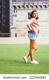 Portrait Of Young American Female College Student In New York City, Wearing Light Pink Leather Jacket, Blue Ripped Denim Shorts, White Sneakers, Hands On Hips, Standing On Green Lawn On Campus.
