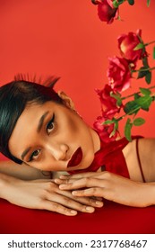 portrait of young and appealing asian fashion model with bold makeup and brunette hair looking at camera while laying on table near roses on red background, stylish spring, fashion shoot - Shutterstock ID 2317768467