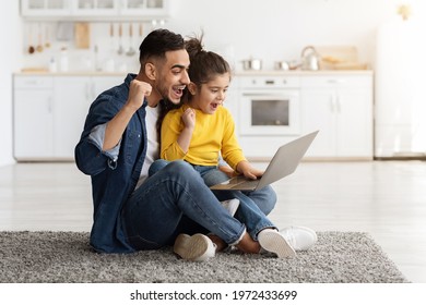 Portrait of young arab dad and little daughter celebrating success with laptop at home, excited happy middle eastern father and small girl looking at computer screen and raising fists with joy
