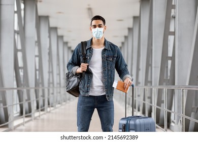 Portrait Of Young Arab Guy Wearing Medical Mask Standing With Luggage At Airport Terminal, Middle Eastern Male Tourist Travelling During Coronavirus Pandemic, Ready For Air Trip, Copy Space