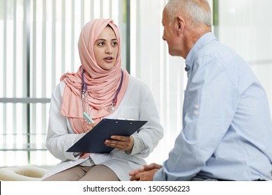 Portrait Of Young Arab Woman Working As Doctor In Medical Clinic And Talking To Senior Patient Filling In Form On Clipboard, Copy Space