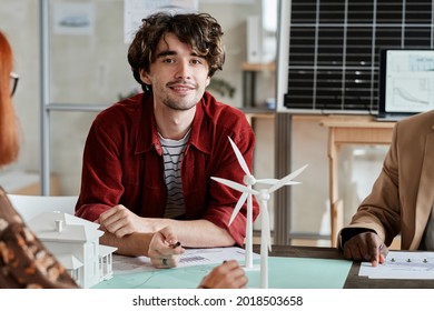 Portrait of young architect smiling at camera while sitting at the table and discussing blueprint with his colleagues at meeting