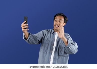 Portrait Of Young Asia Man Making Video Call And Waving At Smartphone Camera Isolated On Blue Background