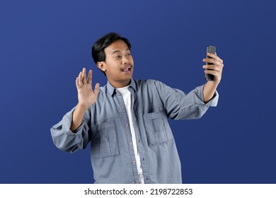 Portrait Of Young Asia Man Making Video Call And Waving At Smartphone Camera Isolated On Blue Background