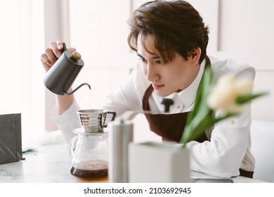 Portrait Of Young Asian Barista Man Wearing Brown Apron Preparing Coffee Using Pour Over Coffee Maker And Drip In Minimal Cafe Shop. Alternative Ways Of Brewing Coffee. Man Barista Cafe Owner Concept