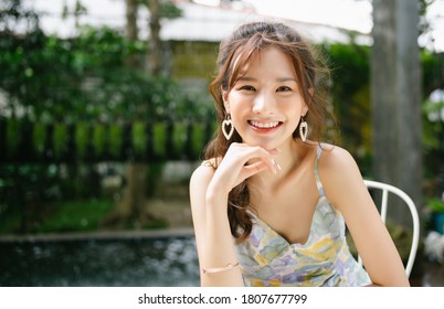 Portrait of young Asian beautiful woman smiling with hand on chin and looking at camera while sitting outdoor on cafe terrace with green plant background. Holiday activity and lifestyle concept.