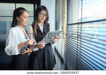 Portrait of young asian busineewoman with glasses holding tablet , smiling and looking outside while standing near window in modern office. Copy space.