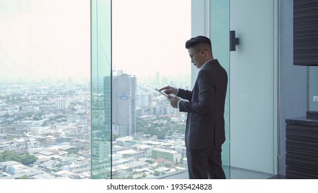 Portrait of young Asian businessman person using a tablet technology device in front of office glass window looking through urban city outside view. People lifestyle.
