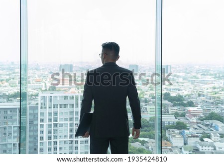 Portrait of young Asian businessman person  thinking, standing in front of office glass window looking through urban city outside view. People lifestyle.
