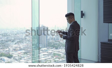 Portrait of young Asian businessman person using a tablet technology device in front of office glass window looking through urban city outside view. People lifestyle.