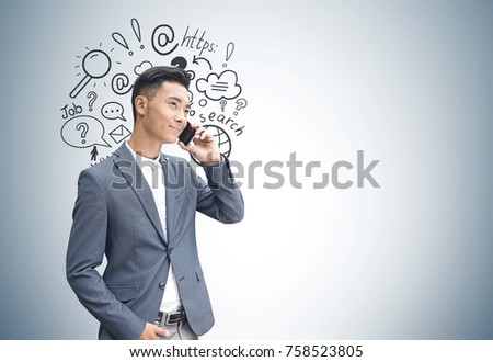 Portrait of a young Asian businessman in a suit talking on the phone and smiling. He is standing near a gray wall an internet search sketch. Mock up