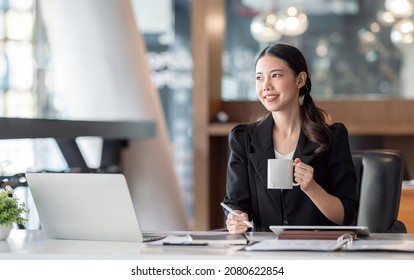 Portrait Of Young Asian Businesswoman In Black Suit Holding Mug And Pen, Smiling And Looking Outside While Sitting In Modern Office Room. Successful Business Concept.