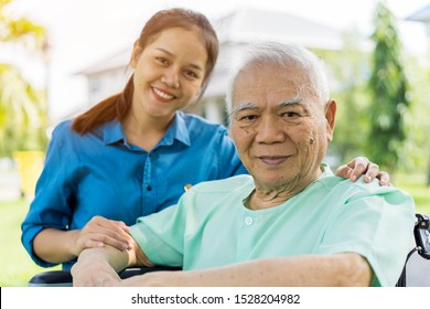 Portrait Of Young Asian Caregiver In Uniform Hugging Smiling Elderly Man Or Patient In Wheelchair During A Home Visit And Spending Time Together. Love, Family Or Assistant Or Elderly Caregiver Concept