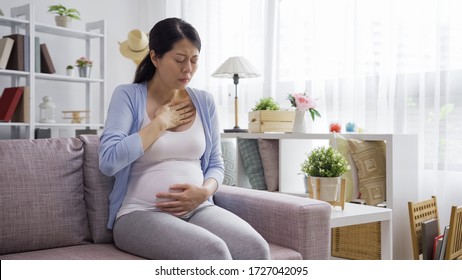 Portrait Of Young Asian Chinese Pregnant Woman Sitting On Couch Hands Touching Heart Feeling Nauseous In 1st Trimester Of Pregnancy. Copy Space. Morning Sickness In Maternal Time Concept.