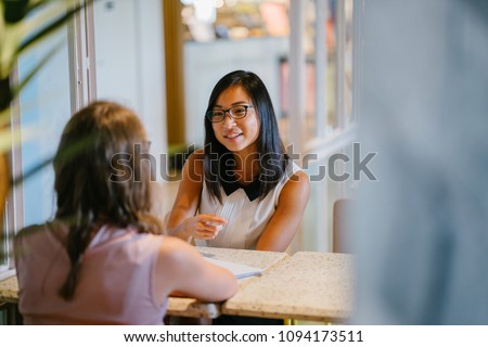 Portrait of a young, Asian Chinese woman in a meeting with a Caucasian woman. She is having a meeting or having an interview and is professionally dressed as she has a discussion at a table indoors. 