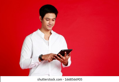 Portrait Young Asian Confident Handsome Man Looking And Working On Digital Tablet While Standing Against Isolated Red Background. Bangkok Thailand South East Asia