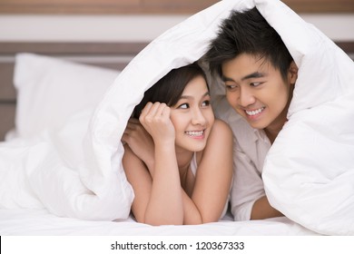 Portrait of young Asian couple looking at one another with smiles under blanket