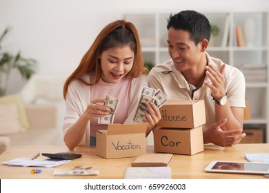 Portrait Of Young Asian Couple Putting Money To Piggy Banks, Saving  For House, Car And Vacation